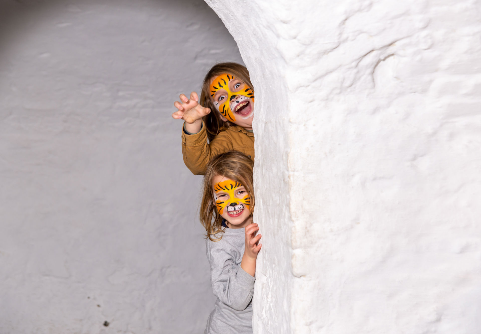 En jente og en gutt med tigermaling i ansiktet titter fram bak en hvit murvegg.