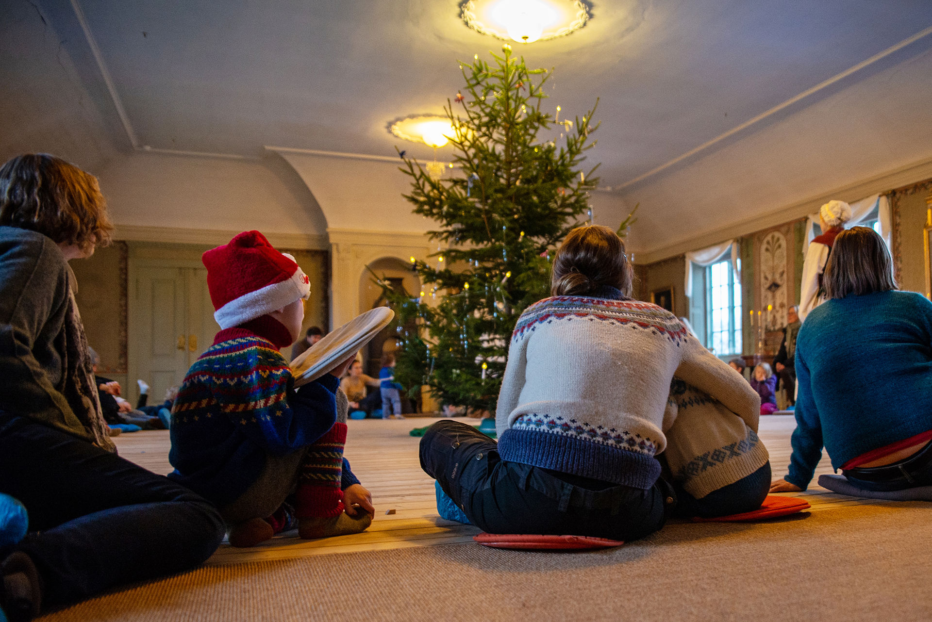 Barn og voksne sitter på gulvet rundt et stort juletre.