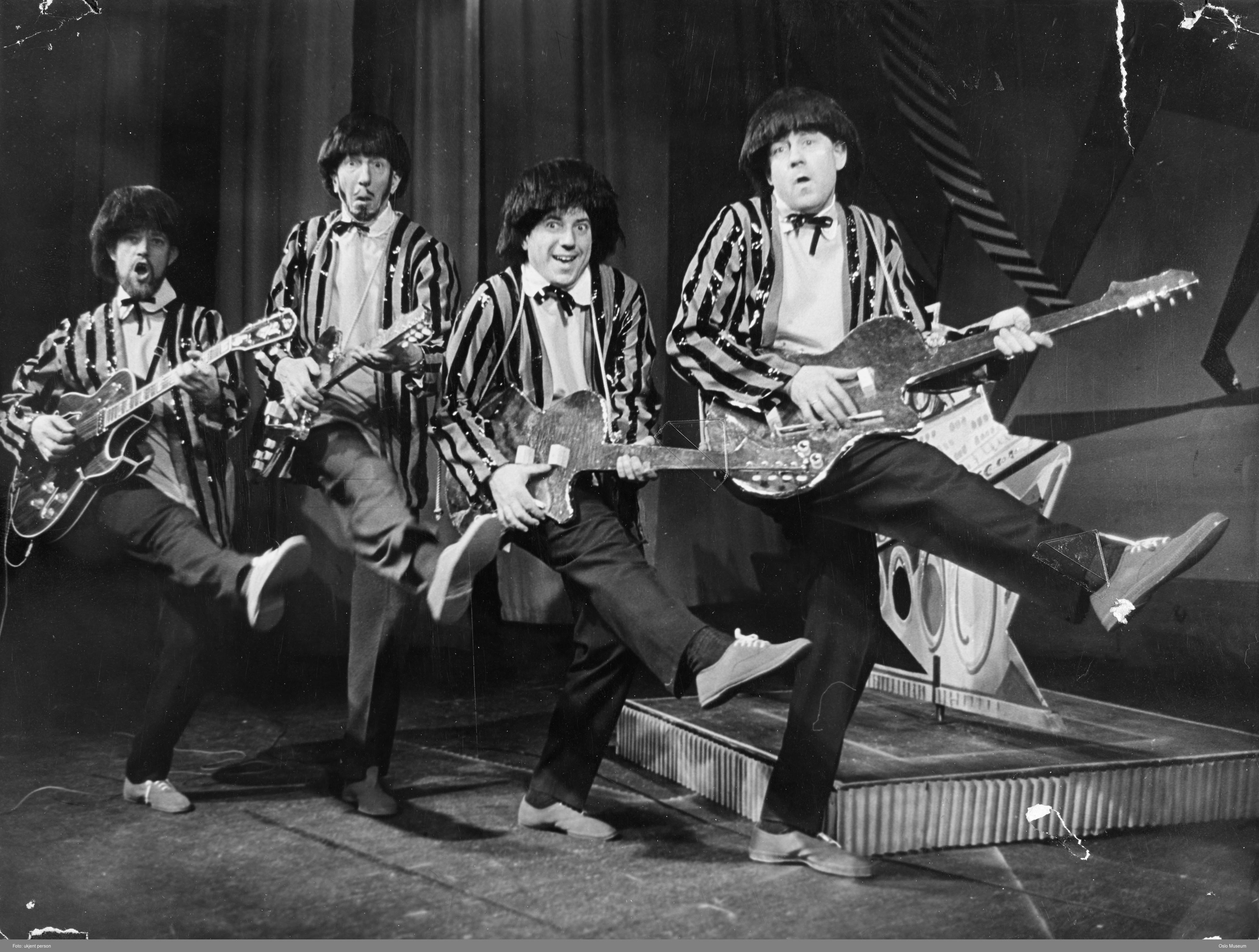 Fire menn i stripete paljettjakker, mørke parykker og med hver sin gitar står på linje med et ben  