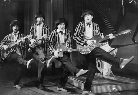 Fire menn i stripete paljettjakker, mørke parykker og med hver sin gitar står på linje med et ben