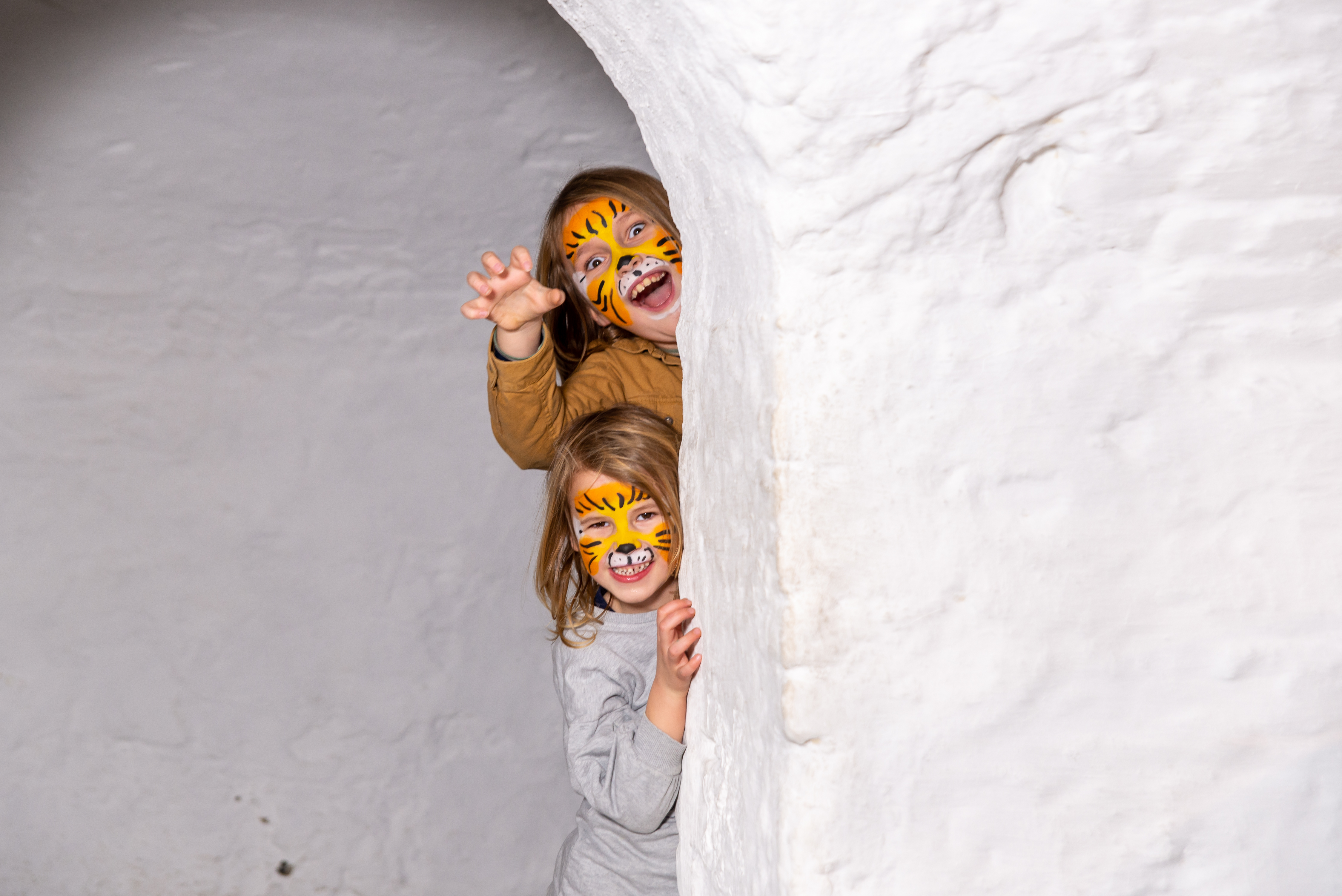 En jente og en gutt med tigermaling i ansiktet titter fram bak en hvit murvegg.