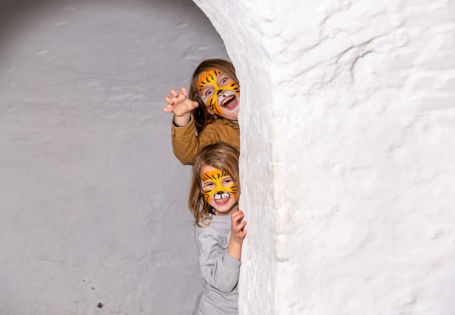 En jente og en gutt med tigermaling i ansiktet titter fram bak en hvit murvegg.