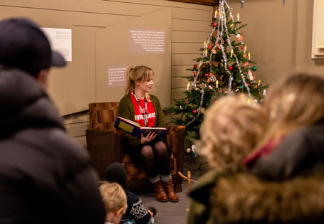 Kvinne sitter i en stol og leser eventyr for barn og voksne som står rundt henne. Et pyntet juletre står ved siden av.