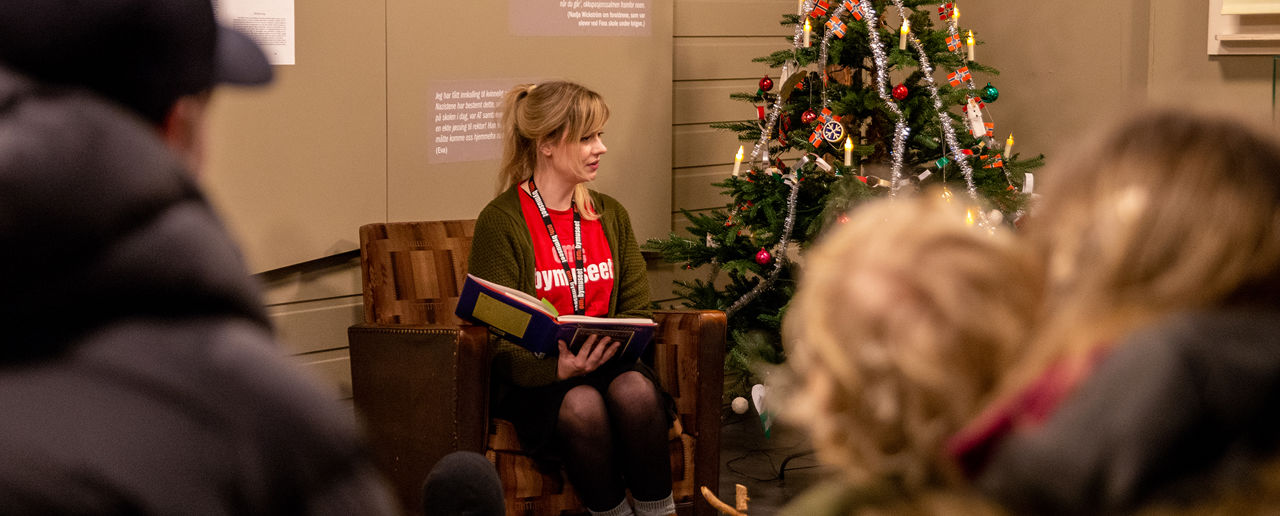 Kvinne sitter i en stol og leser eventyr for barn og voksne som står rundt henne. Et pyntet juletre står ved siden av.