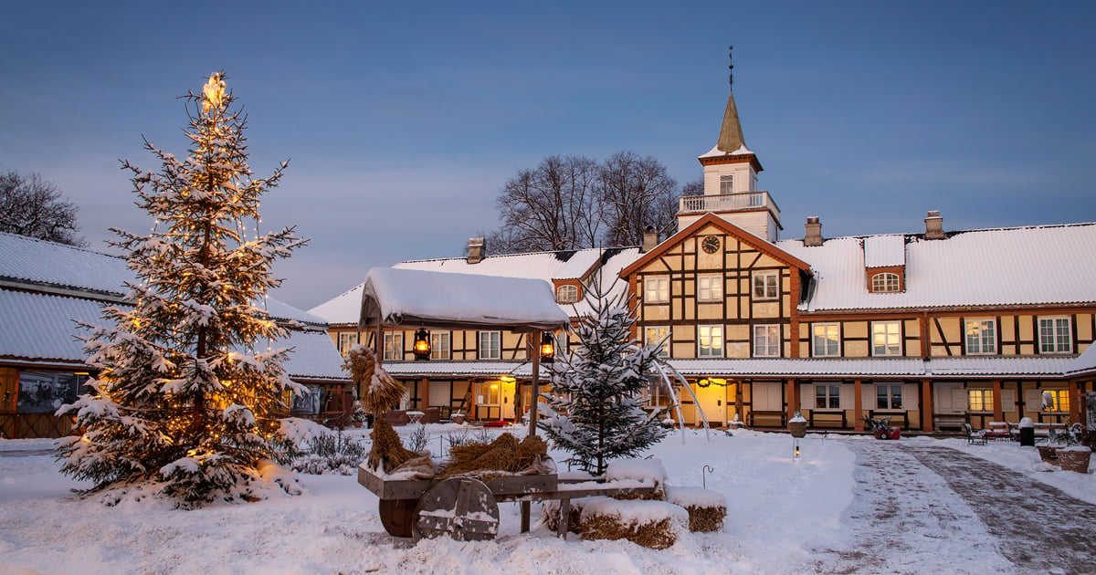Gammel herregård med stort tun foran. Hvit snø på taket og på tunet foran. Juletre og vogn med julenek i forgrunnen.