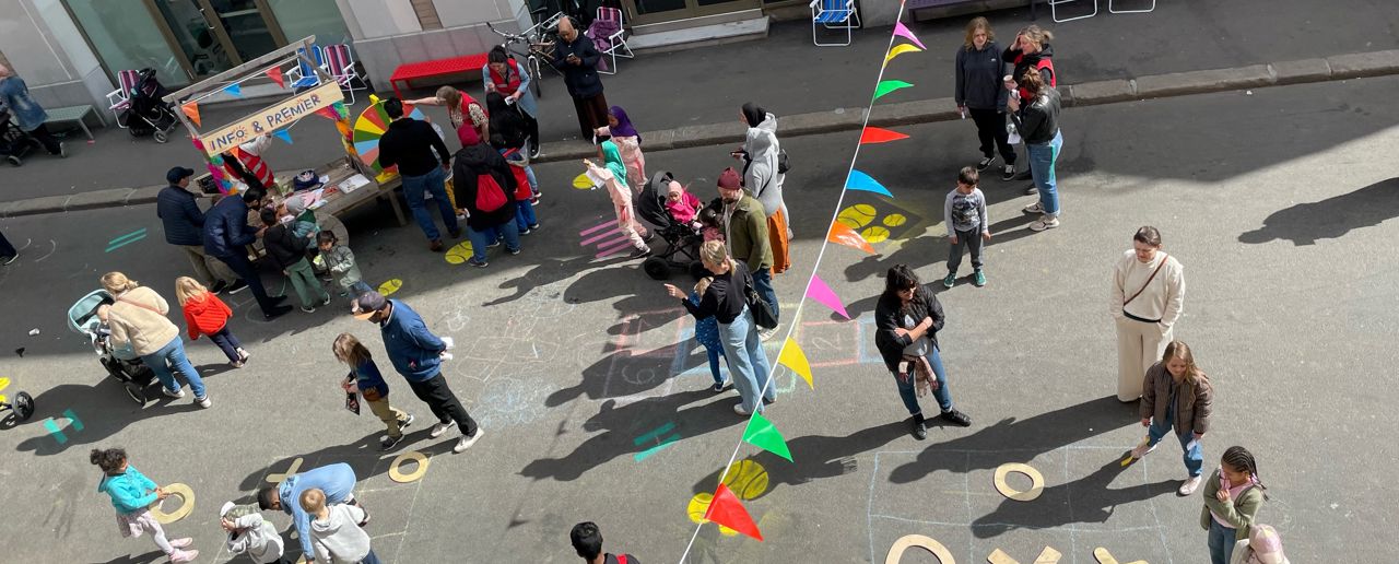 Barn og voksne leker i gaten foran Interkulturelt Museum. Fargerike vimpler henger over gaten, det er kritt-tegninger på asfalten.
