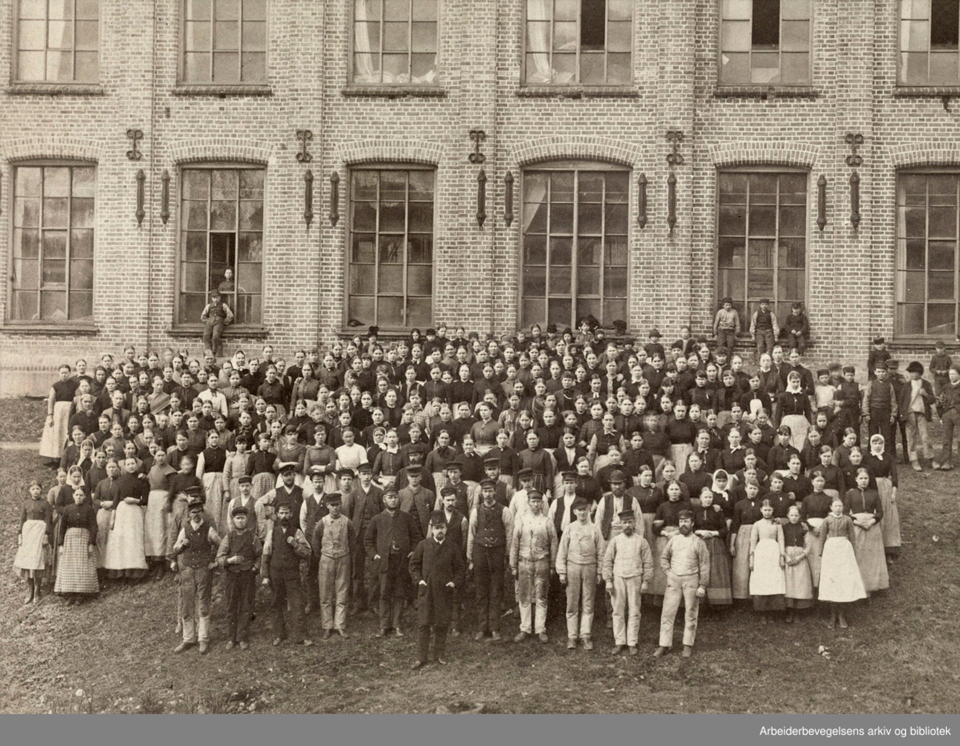 Sort-hvitt-foto av mange mennesker i gammeldagse arbeidsklær oppstilt utenfor et murbygg