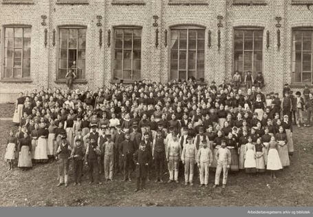 Sort-hvitt-foto av mange mennesker i gammeldagse arbeidsklær oppstilt utenfor et murbygg