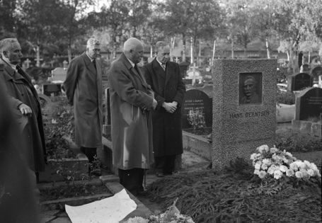 Sorthvitt-bilde av fire menn i frakk som ser ned på en blomsterdekket grav med gravstenen til Hans Berntsen