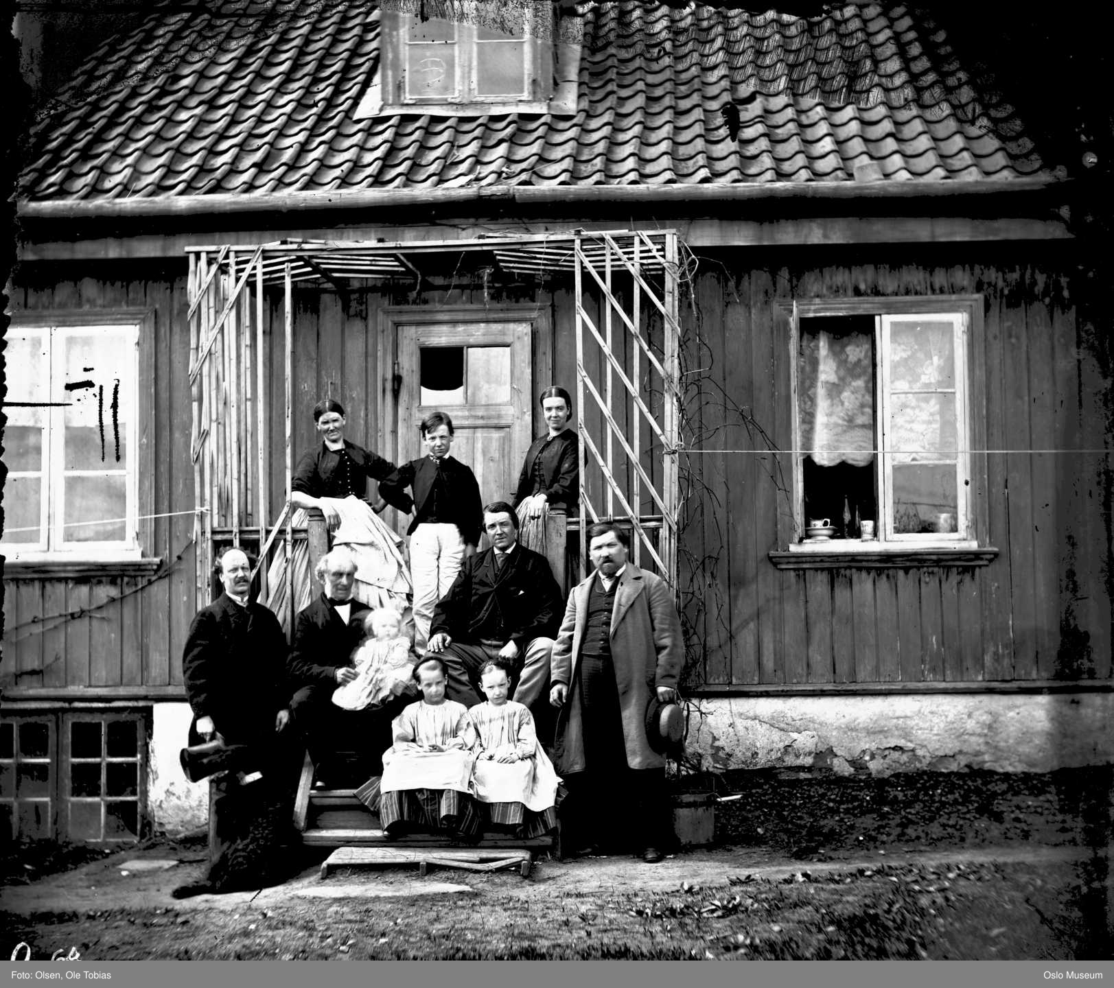 sorthvitt av voksne og barn samlet på en trapp foran et gammelt litt slitent trehus