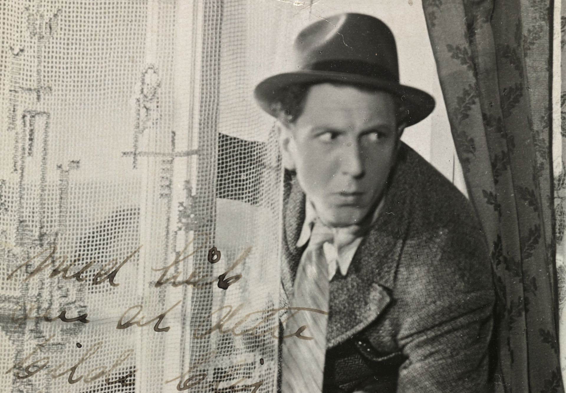 Sorthvitt-bilde av Leif Juster ikledd hatt, frakk og slips på vei inn gjennom et vindu