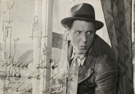 Sorthvitt-bilde av Leif Juster ikledd hatt, frakk og slips på vei inn gjennom et vindu