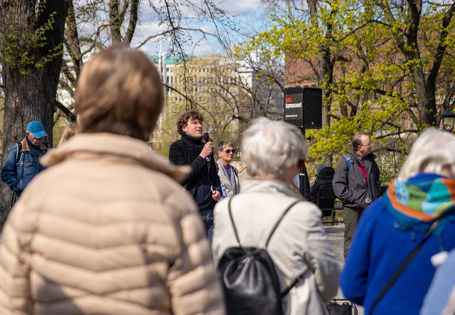 Mann med mikrofon og høyttaler snakker til mennesker. Mannen er vendt mot kamera, publikum ser vi kun ryggen til.