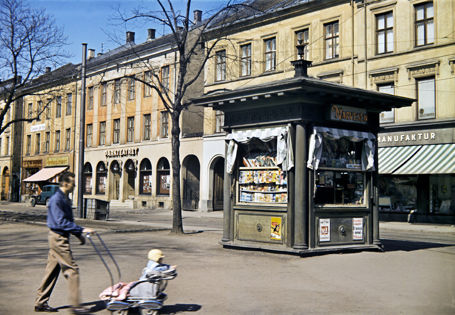 Mann triller baby i en gammeldags barnevogn. I bakgrunnen en frittstående Narvesenkiosk.