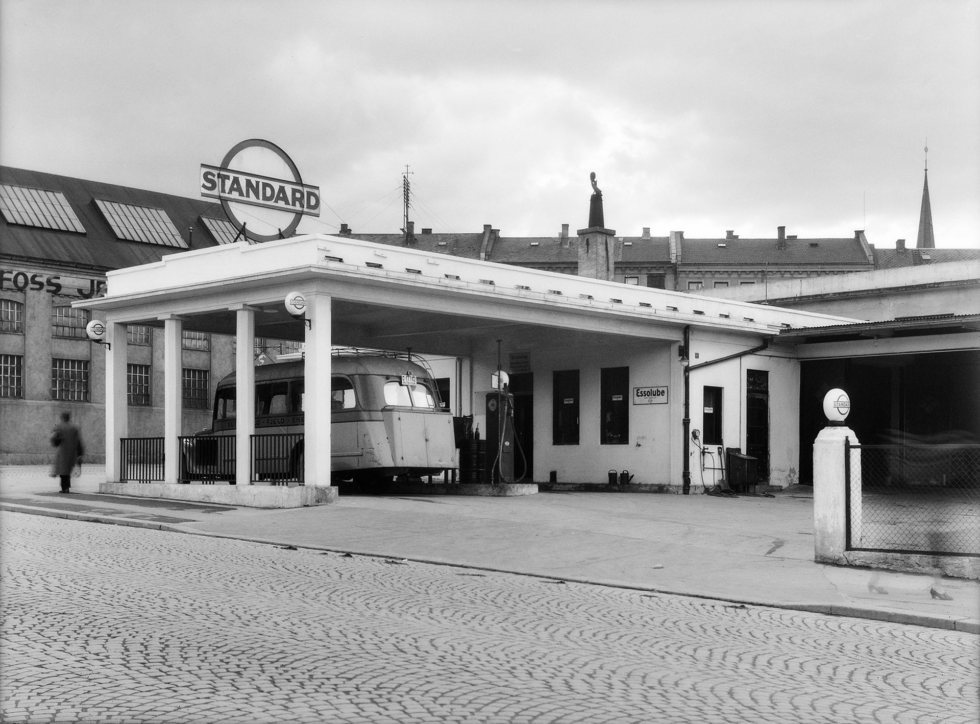 En gammeldags kort buss står parkert under tak på en bensinstasjon.