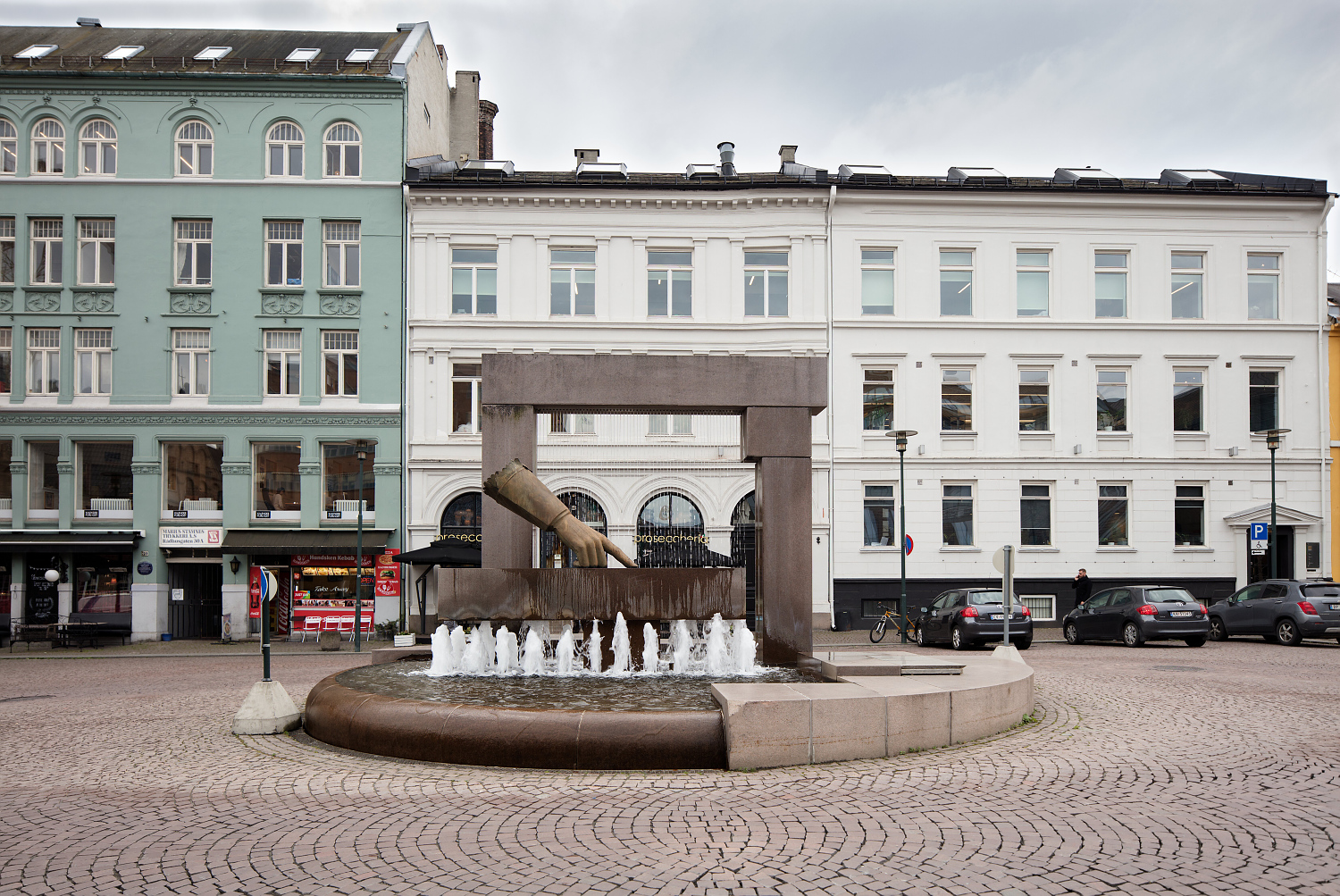 fontene og skulptur av en hånd som peker nedover midt på en brosteinlagt plass