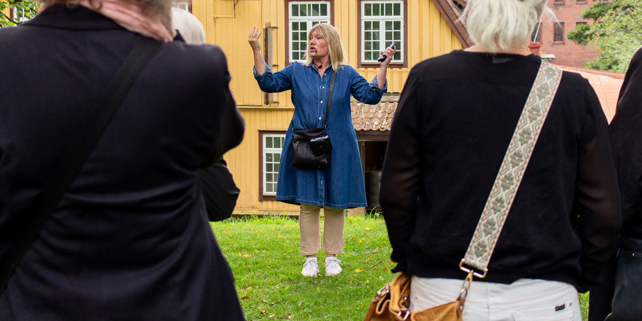 En guide snakker til tilhørere. I bakgrunnen et stort gult trebygg.