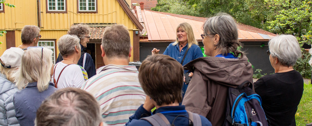 En guide snakker til tilhørere. I bakgrunnen et stort gult trebygg.