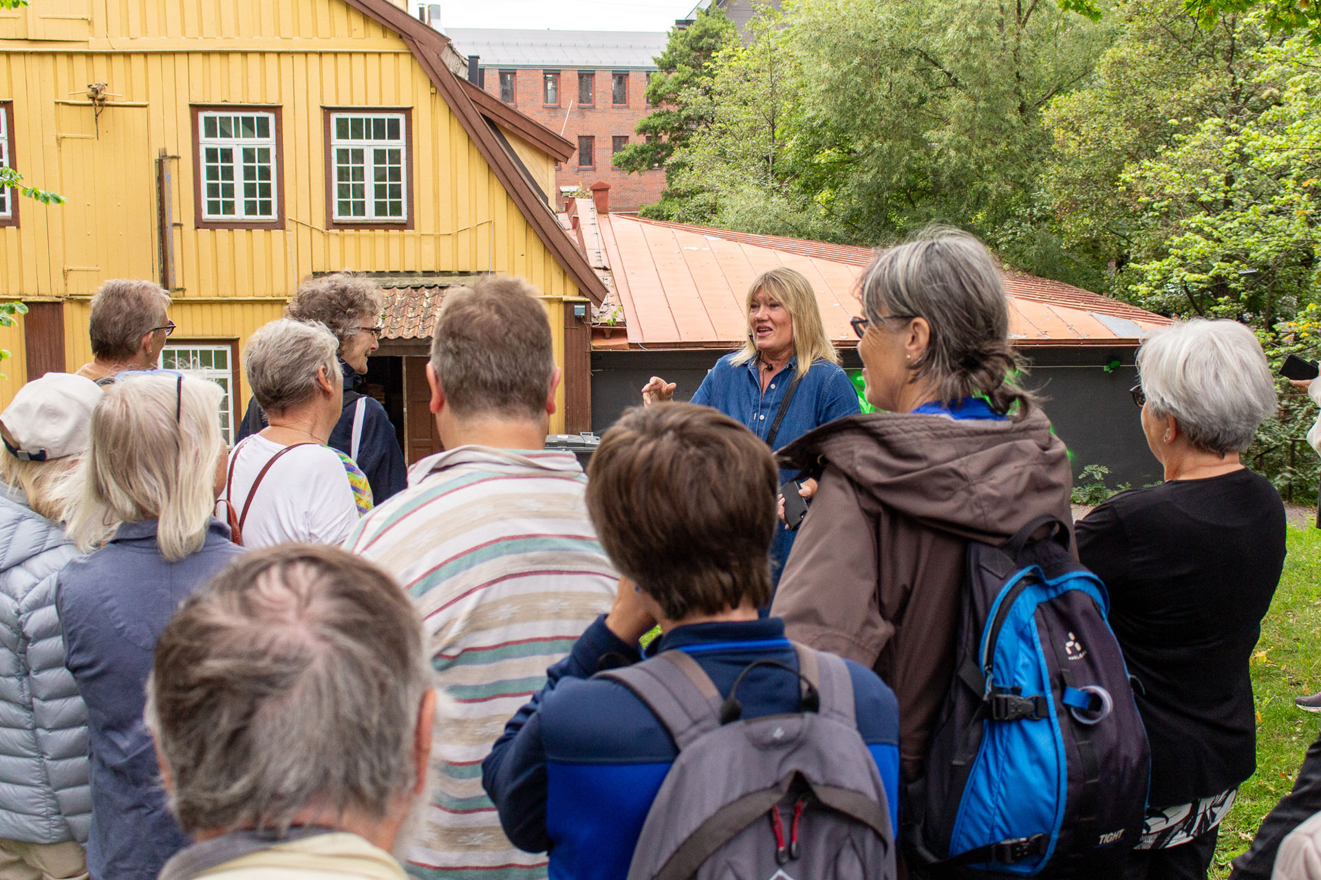 En guide snakker til tilhørere. I bakgrunnen et stort gult trebygg.