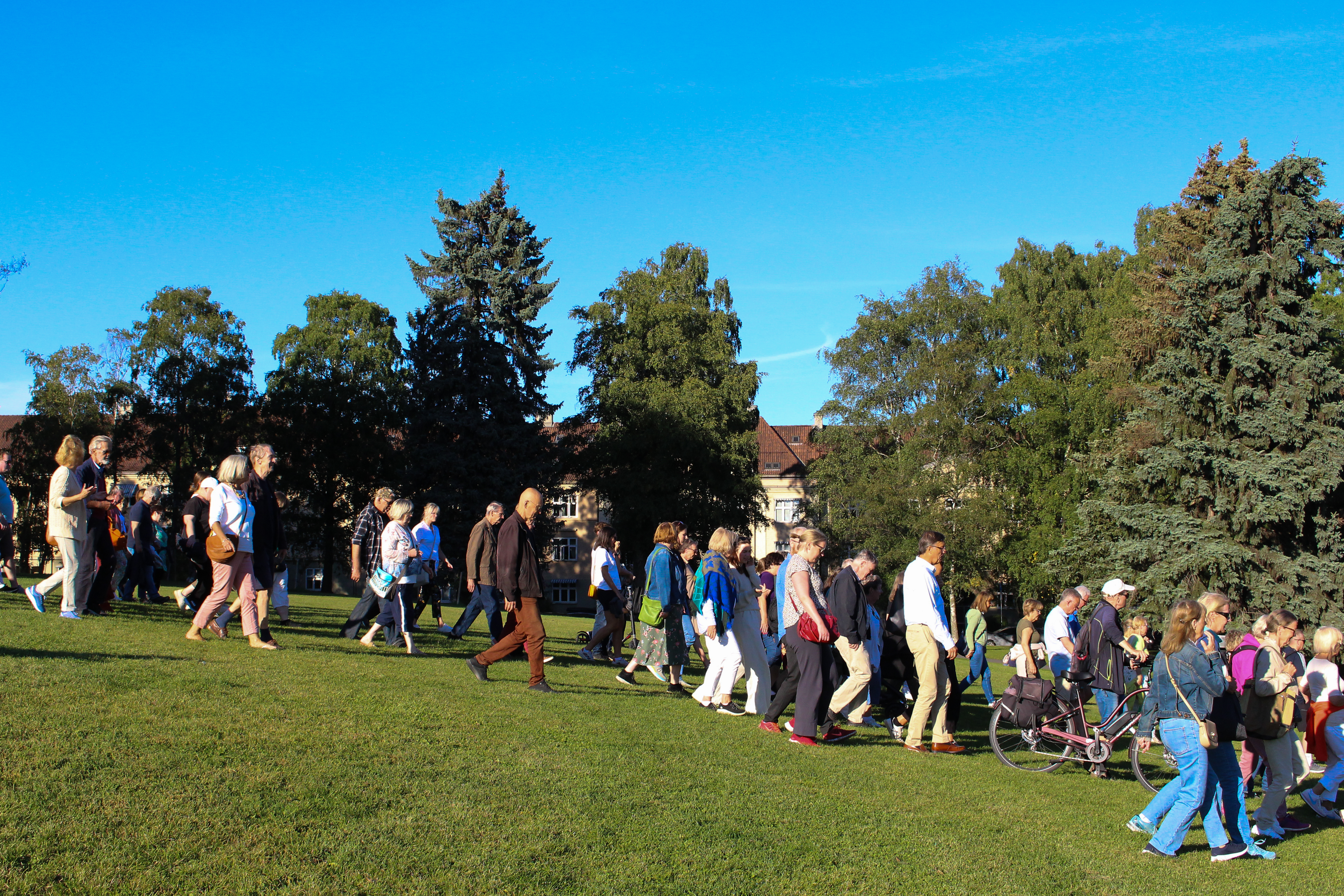 Mange personer går nedover en grønn slette med trær og blå himmel i bakgrunnen