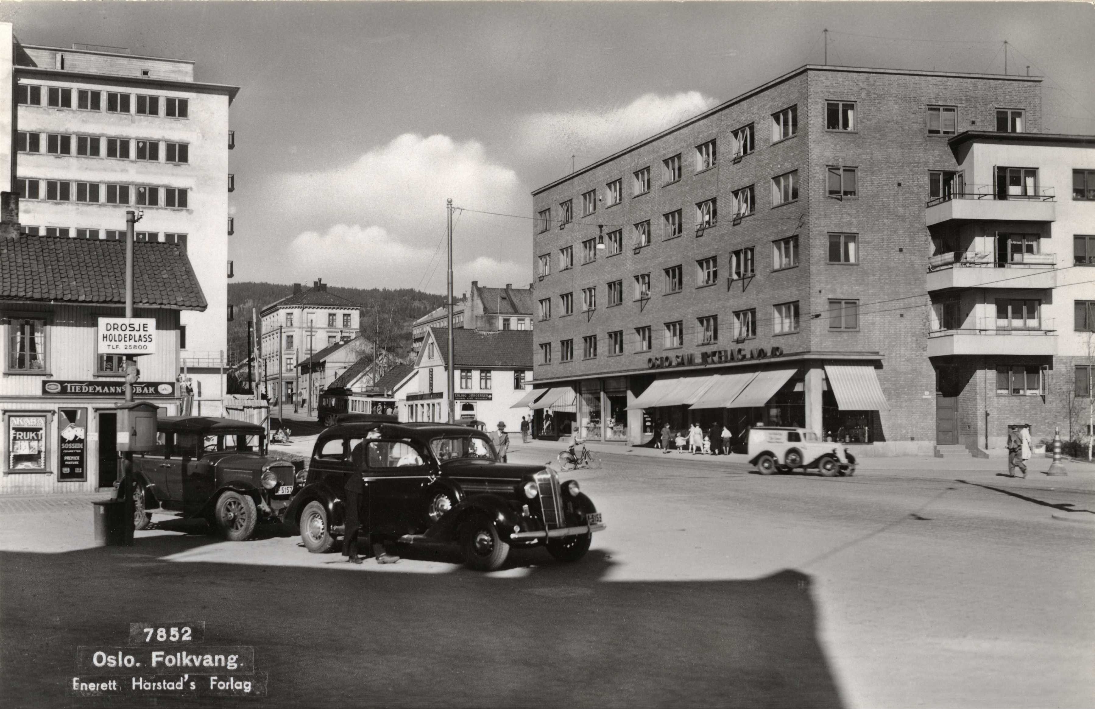 sort-hvitt av to gamle biler på et torv med bolig- og butikkbebyggelse rundt