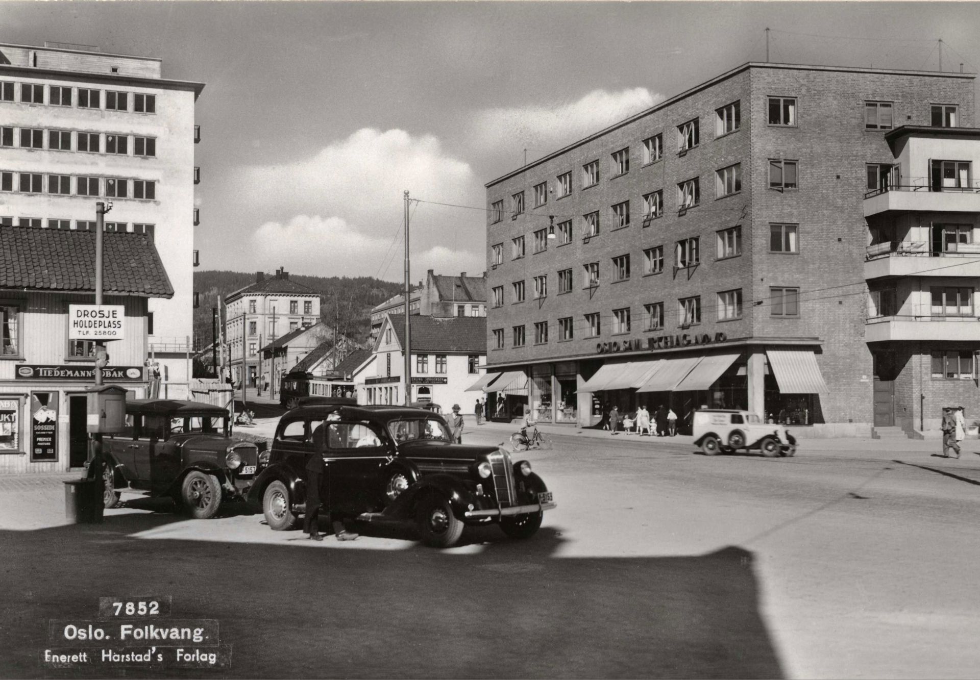 sort-hvitt av to gamle biler på et torv med bolig- og butikkbebyggelse rundt