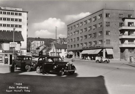 sort-hvitt av to gamle biler på et torv med bolig- og butikkbebyggelse rundt