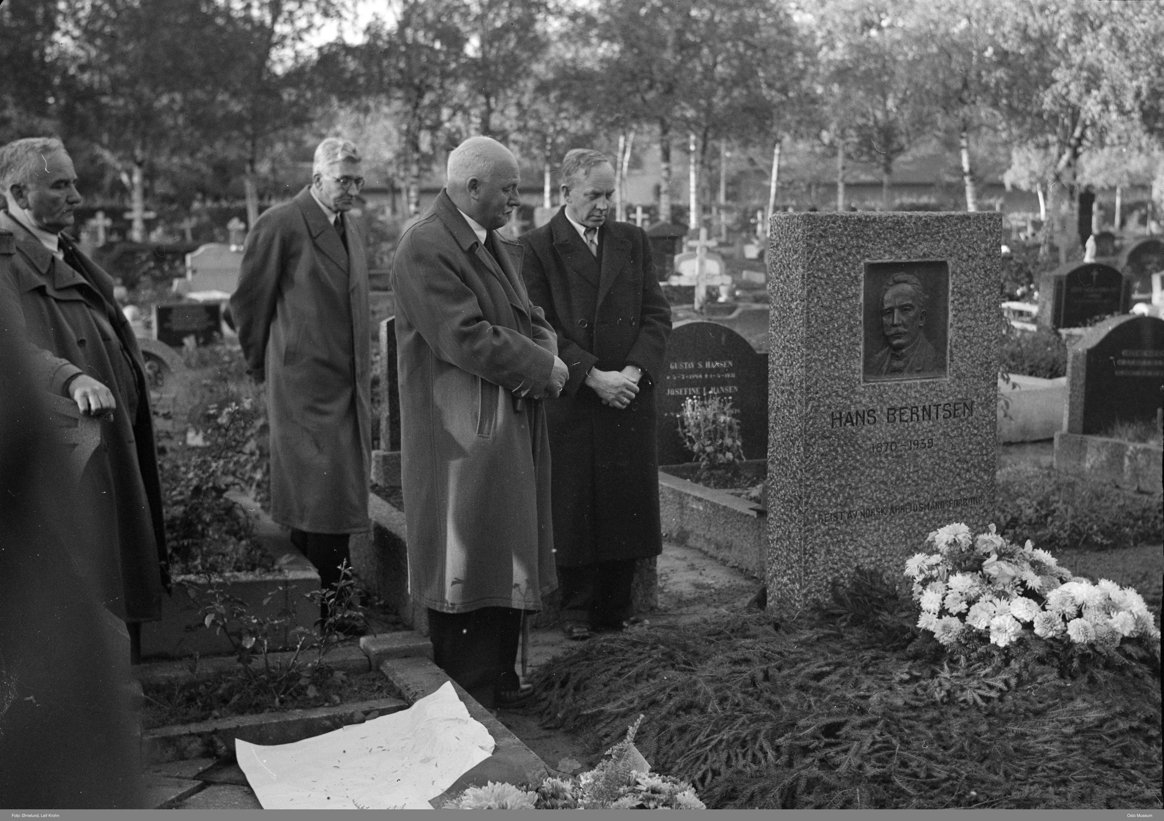 Sorthvitt-bilde av fire menn i frakk som ser ned på en blomsterdekket grav med gravstenen til Hans Berntsen