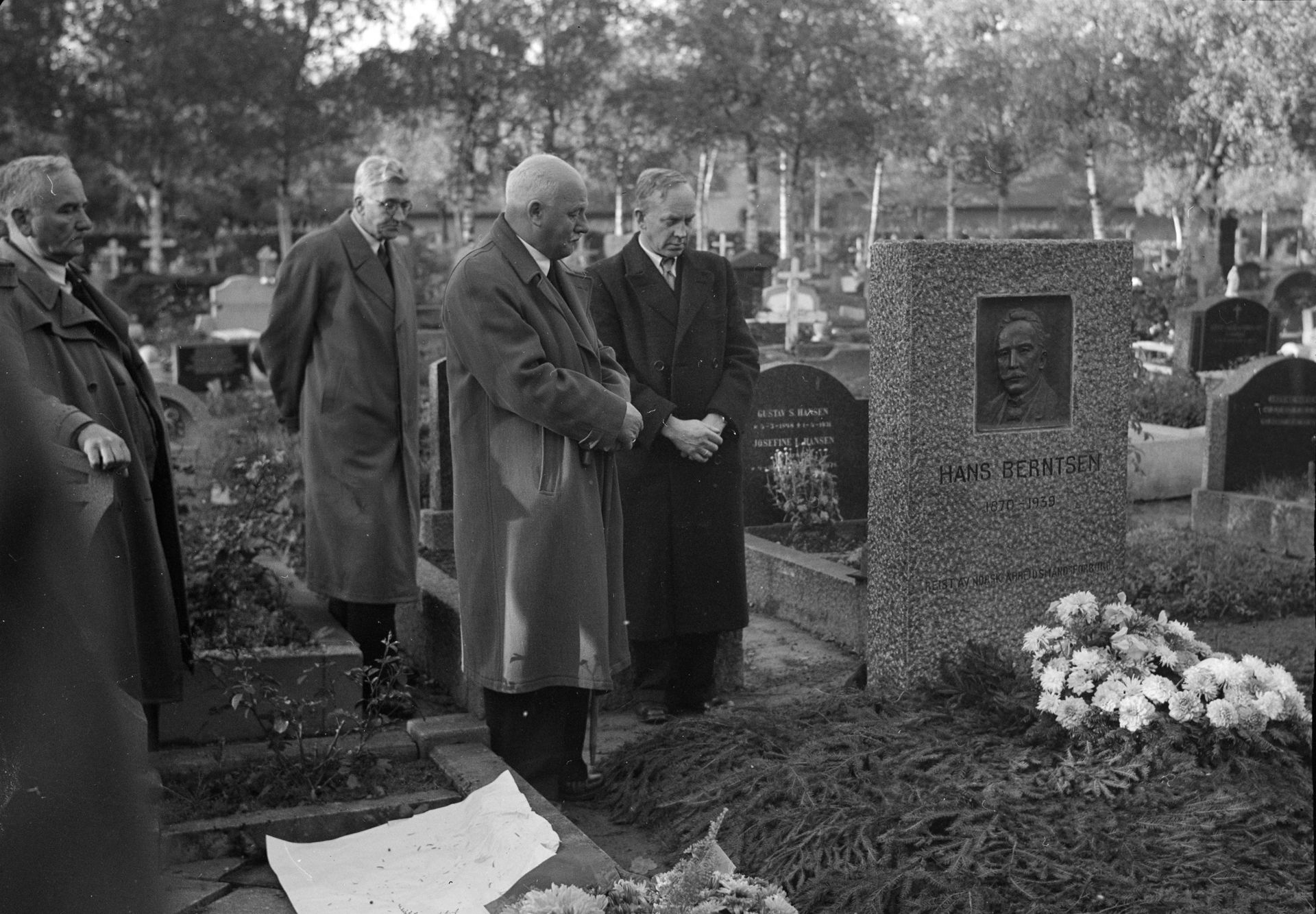 Sorthvitt-bilde av fire menn i frakk som ser ned på en blomsterdekket grav med gravstenen til Hans Berntsen