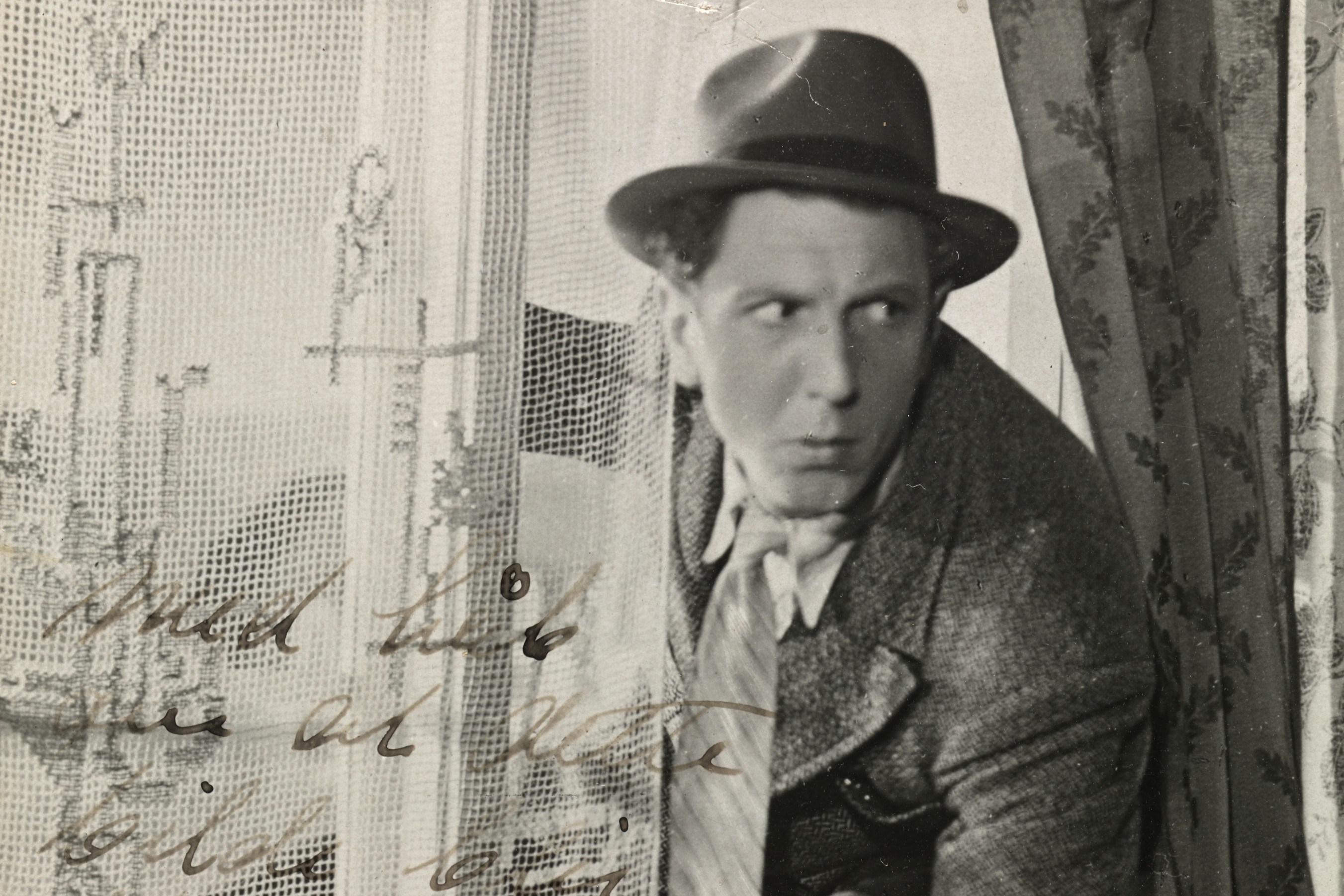 Sorthvitt-bilde av Leif Juster ikledd hatt, frakk og slips på vei inn gjennom et vindu