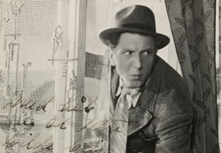 Sorthvitt-bilde av Leif Juster ikledd hatt, frakk og slips på vei inn gjennom et vindu