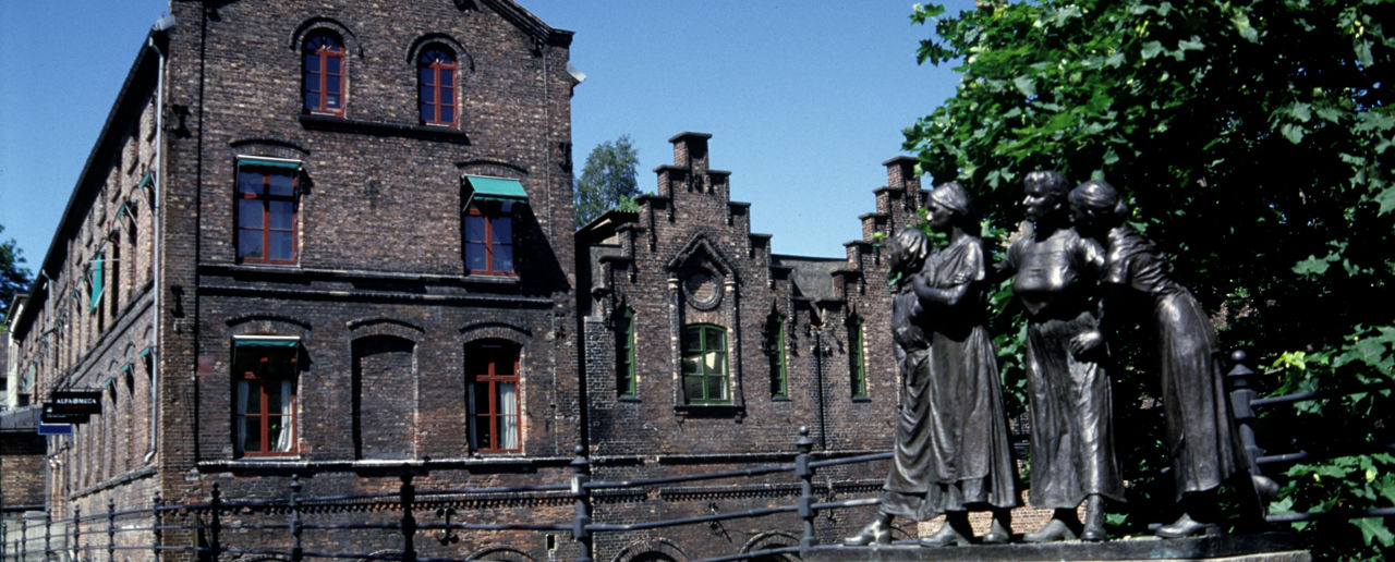 Stort murbygg i bakgrunnen og skulptur av fire arbeiderjenter