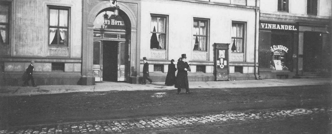 Sort-hvitt-bilde av en frakkledd Henrik Ibsen i flosshatt utenfor Grand Hotel