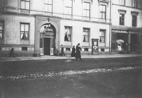 Sort-hvitt-bilde av en frakkledd Henrik Ibsen i flosshatt utenfor Grand Hotel