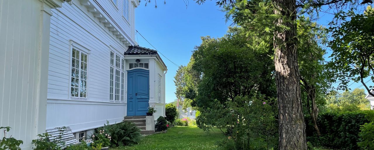 Hvitt trehus med blå inngangsdør og grønn hage rundt