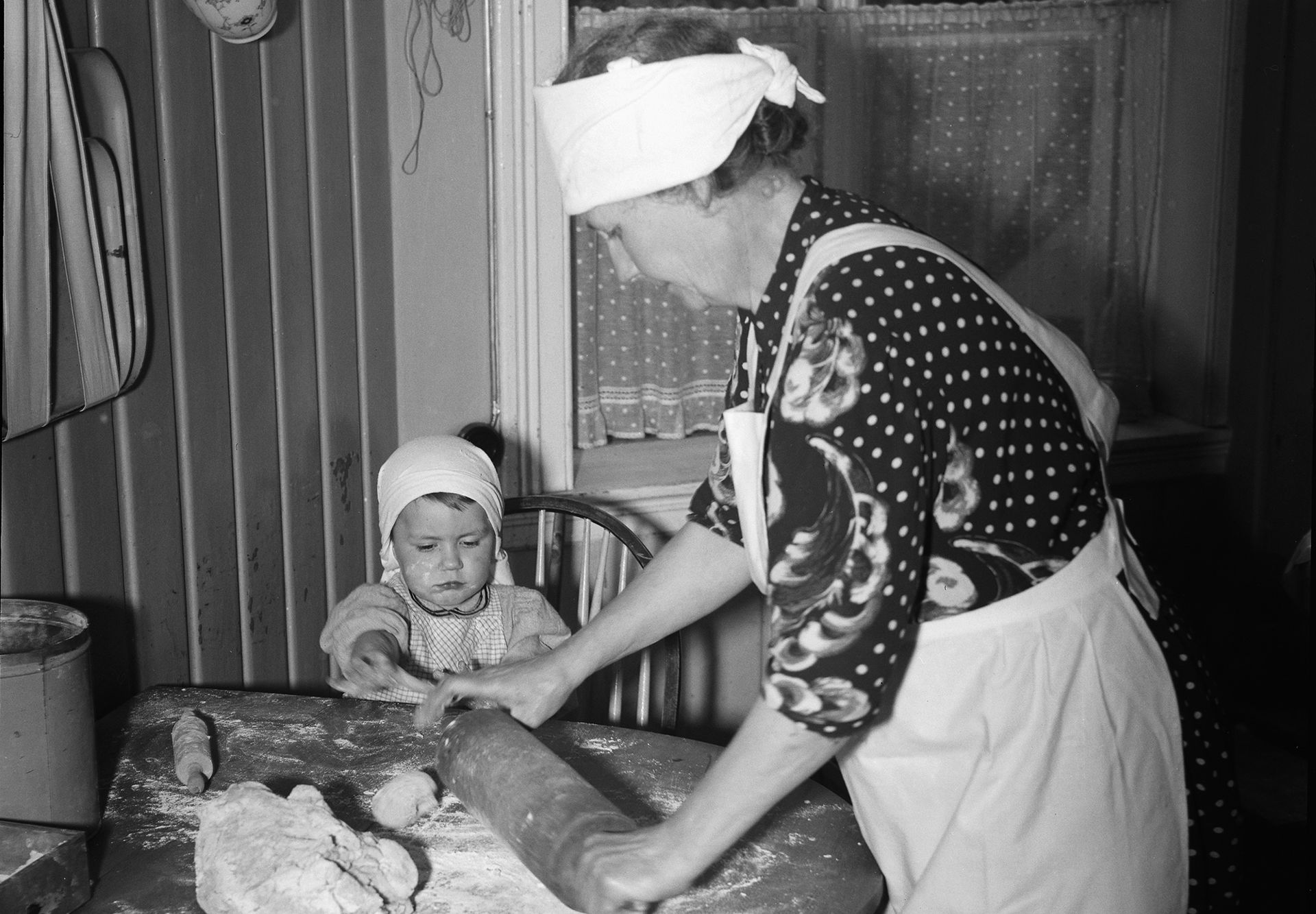 Bestemor bruker kjevle på en deig, et lite barn ser på. Sort-hvitt bilde.