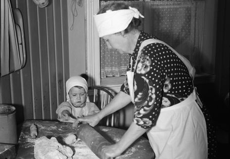 Bestemor bruker kjevle på en deig, et lite barn ser på. Sort-hvitt bilde.