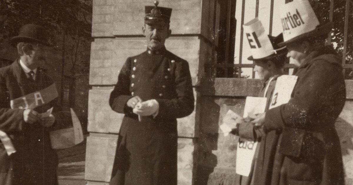 Tre personer står utenfor en bygning, med en uniformert politimann i midten og personer med plakater på hver side