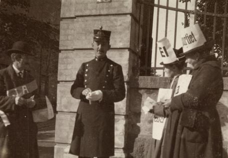 Tre personer står utenfor en bygning, med en uniformert politimann i midten og personer med plakater på hver side
