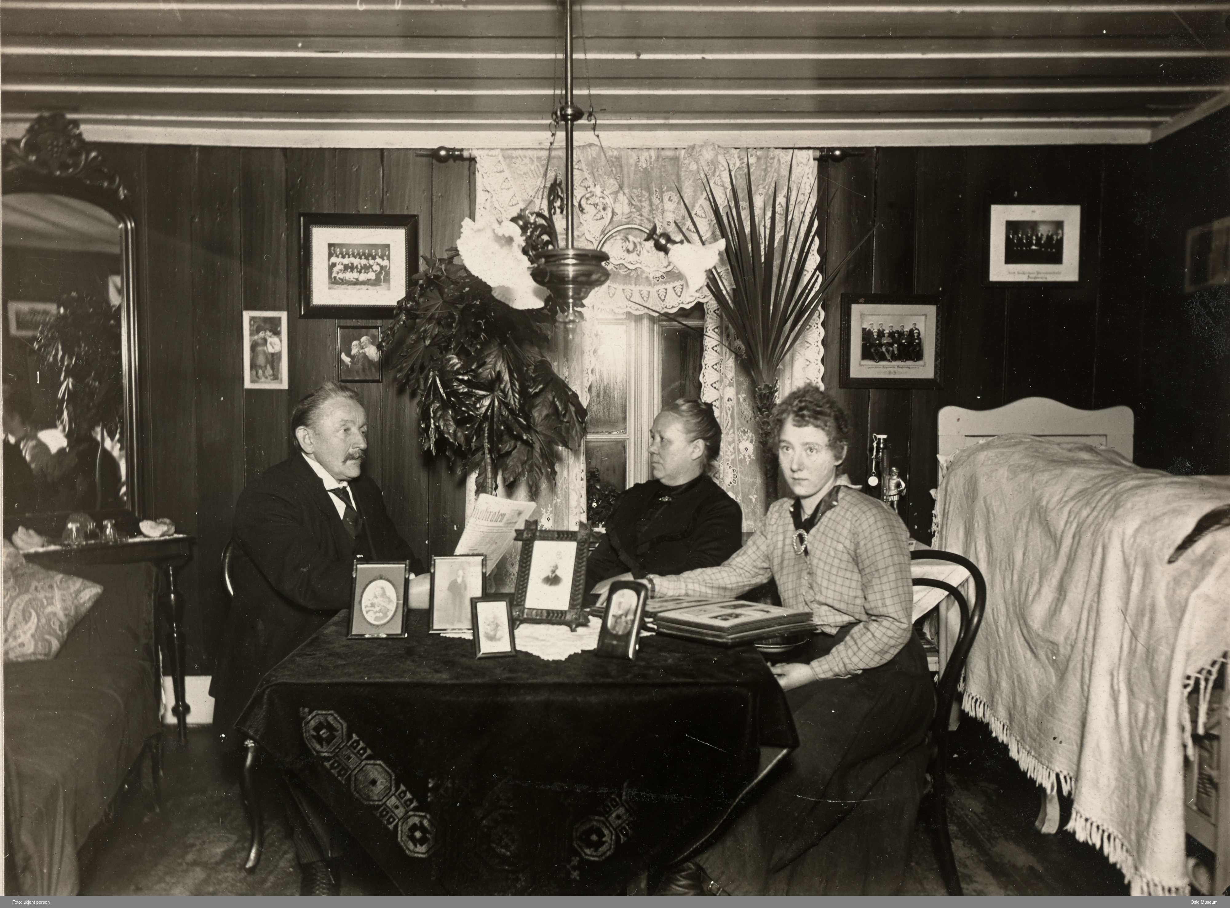 Sort-hvitt gammelt bilde av en mann og to kvinner i en gammeldags stue rundt et rundt bord.