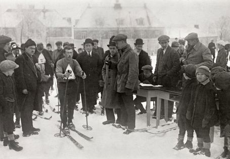 Mann med ski på beina og startnummer på brystet er omringet av mange mennesker i vinterklær.