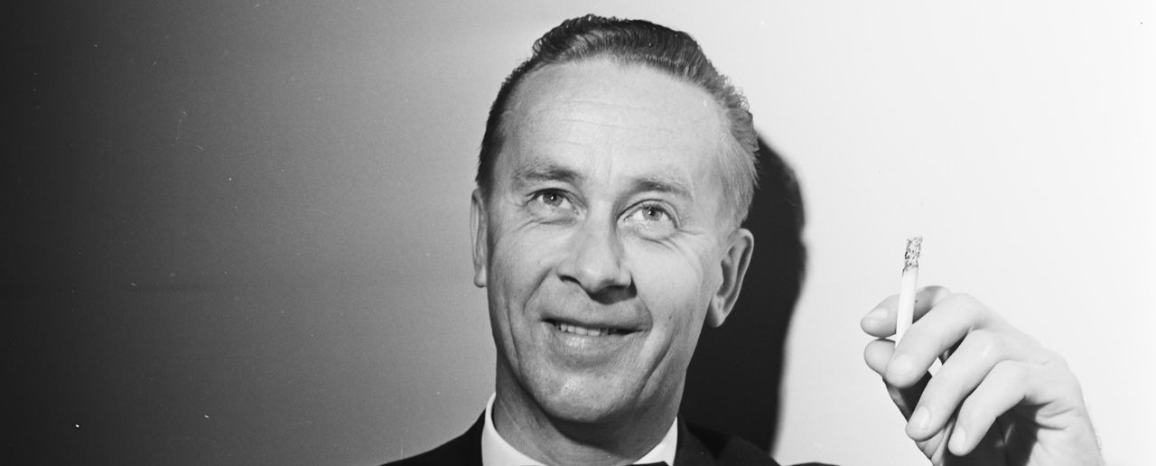 Smilende mann i sort dress fotografert i sort-hvitt. Sigarett i hånden.
