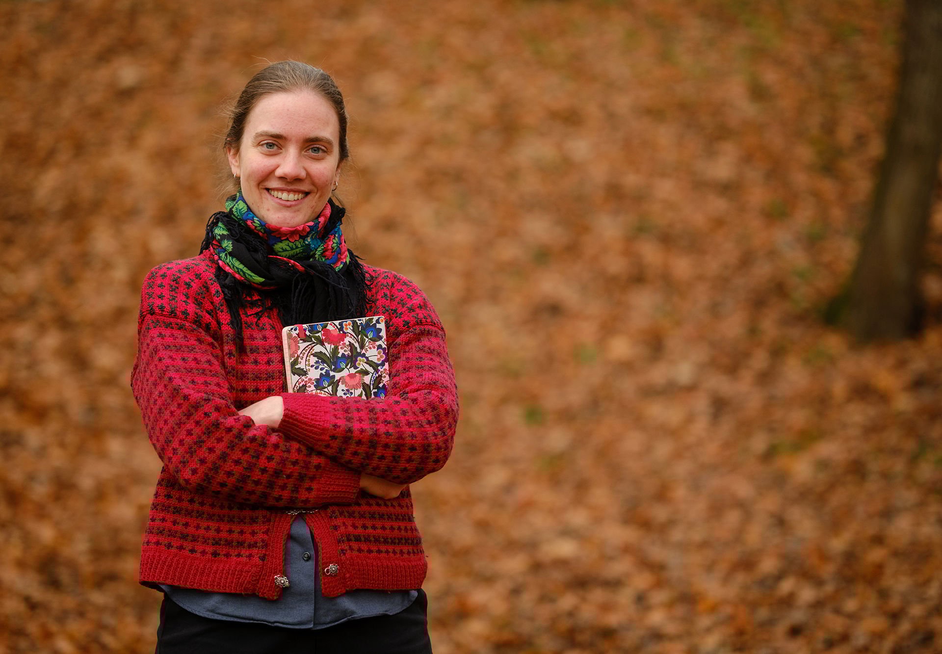 Kvinne i rød strikkegenser og fargerikt skjerf smiler mot kamera med foldede armer med en bok i armene