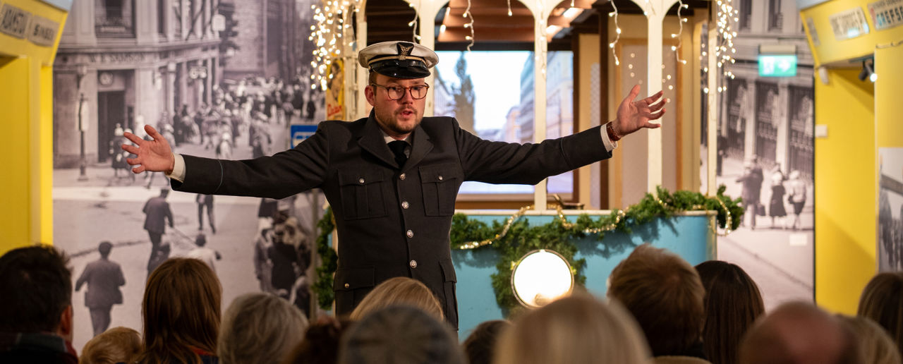 Mann i trikkeuniform med hvit lue og sort dress står med armene ut foran en blå trikk i en utstilling