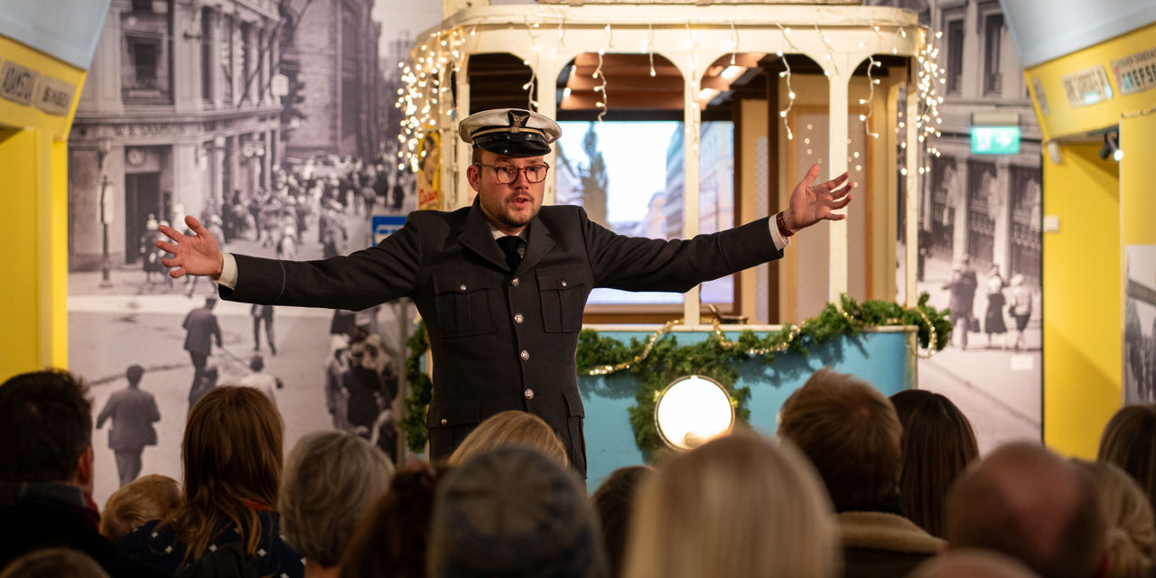 Mann i trikkeuniform med hvit lue og sort dress står med armene ut foran en blå trikk i en utstilling