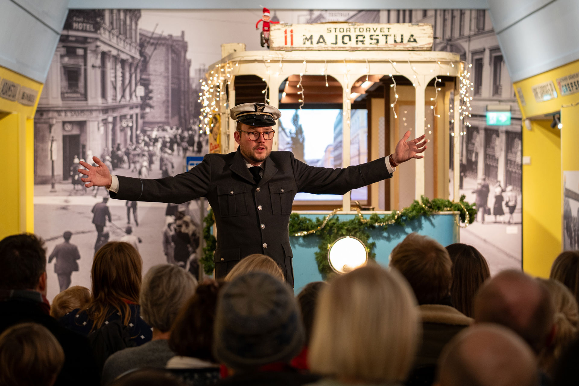 Mann i trikkeuniform med hvit lue og sort dress står med armene ut foran en blå trikk i en utstilling