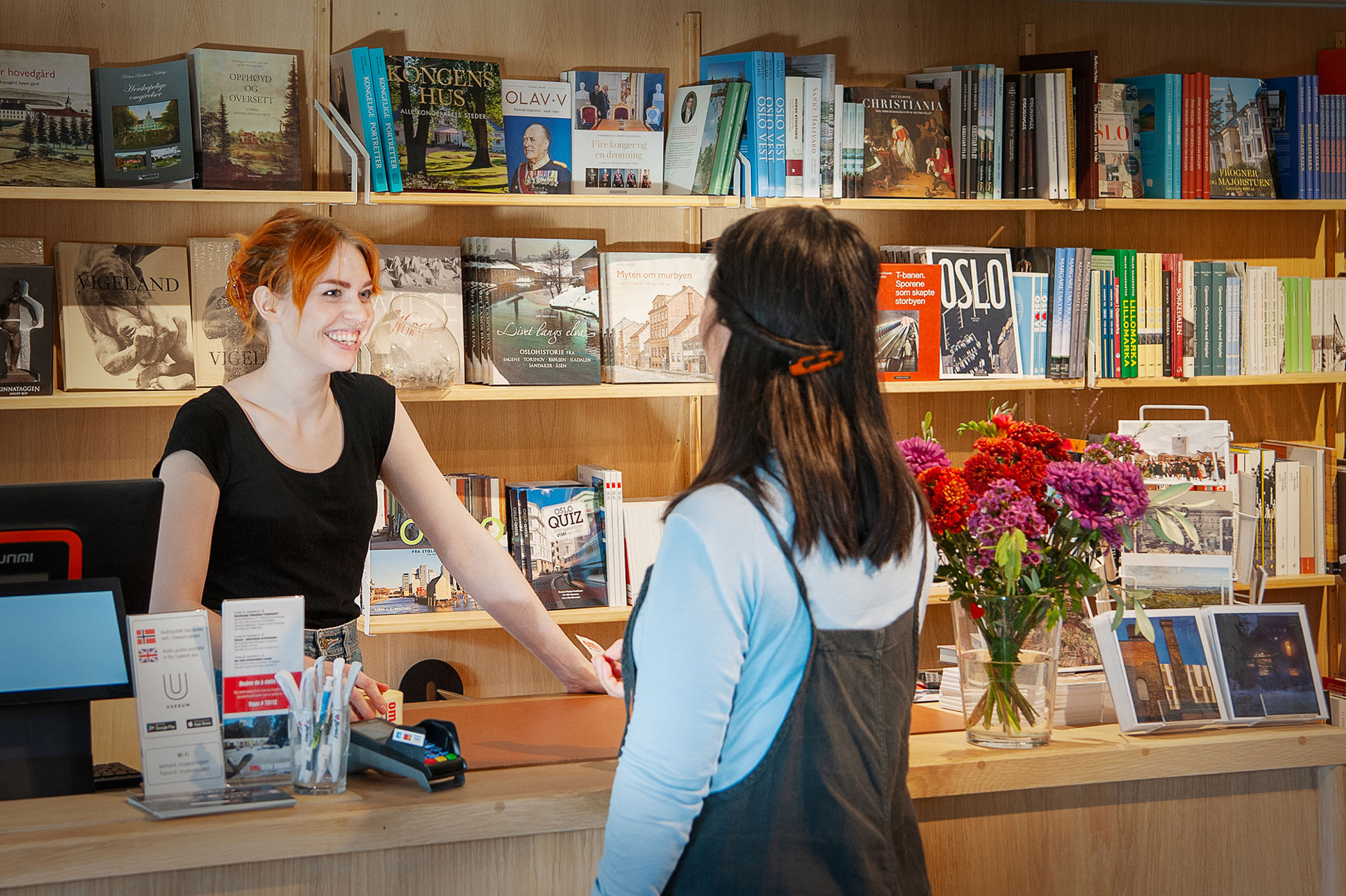 Ung kvinne bak kassen betjener kvinnelig kunde i butikken. Masse bøker i bakgrunnen.