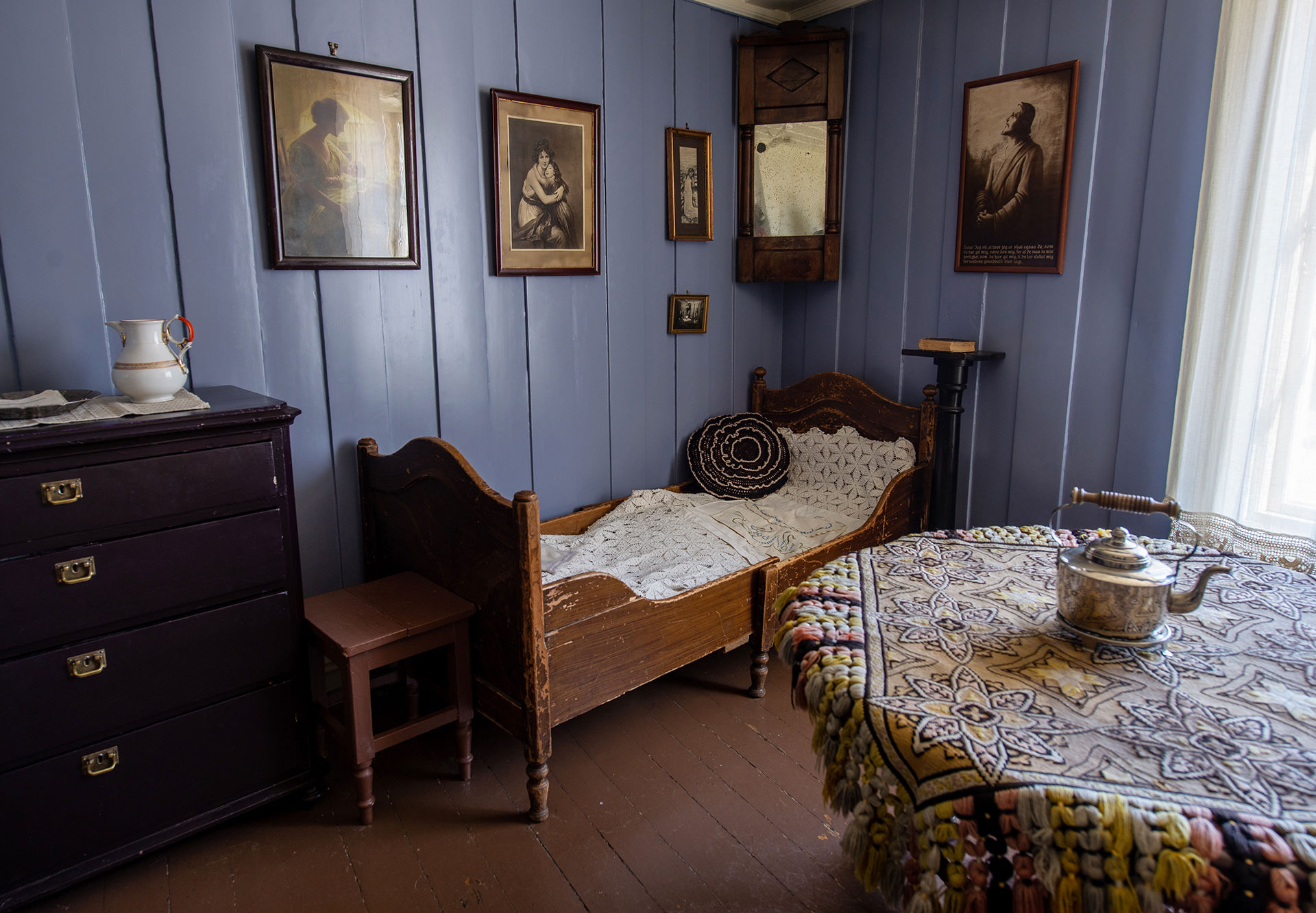 Kommode, gammel seng og et bord med en tekanne på. Det henger bilder og malerier på den blå veggen.