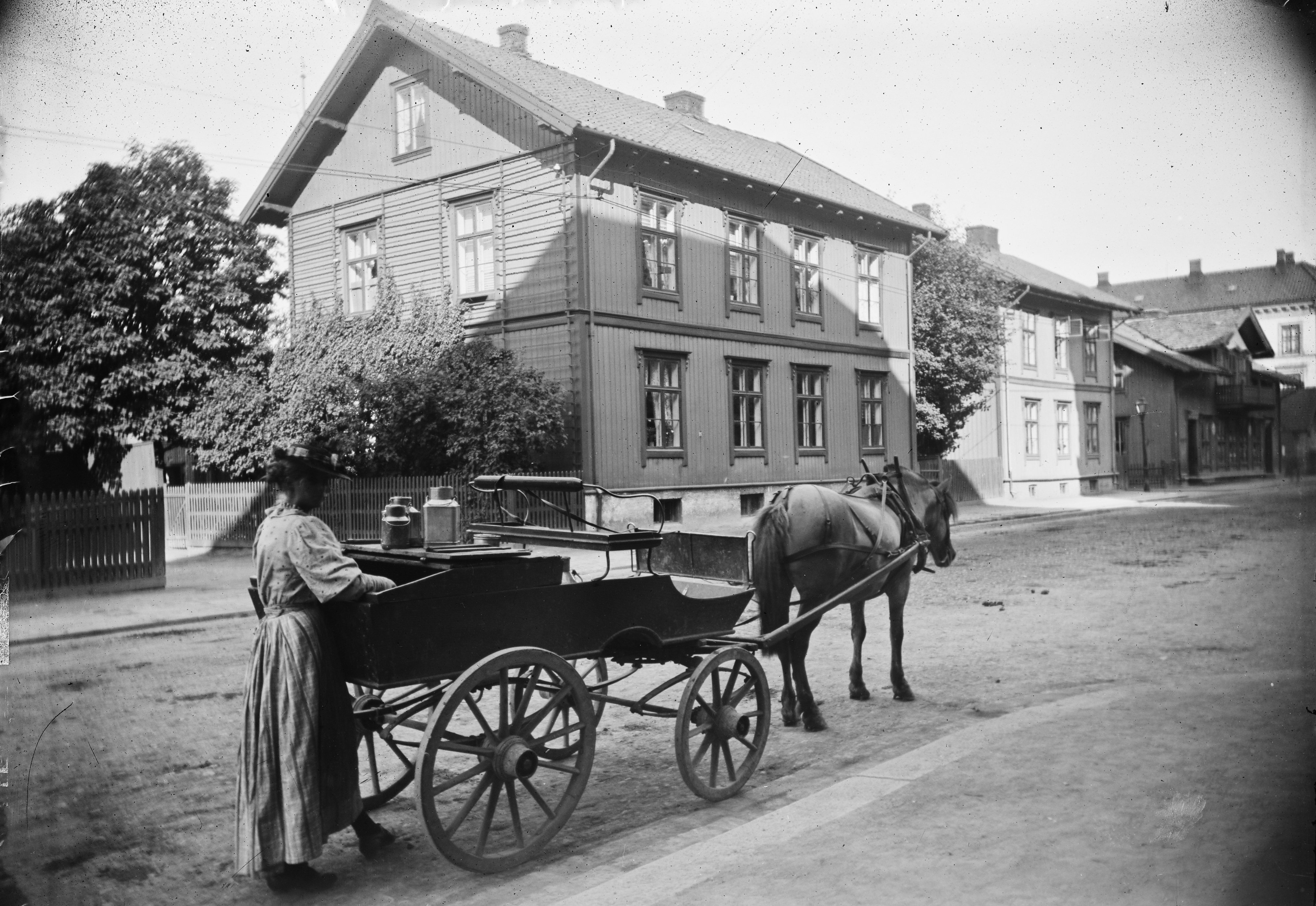 Gate med hus og i en kvinne som st&aring;r ved en hest med melkevogn 