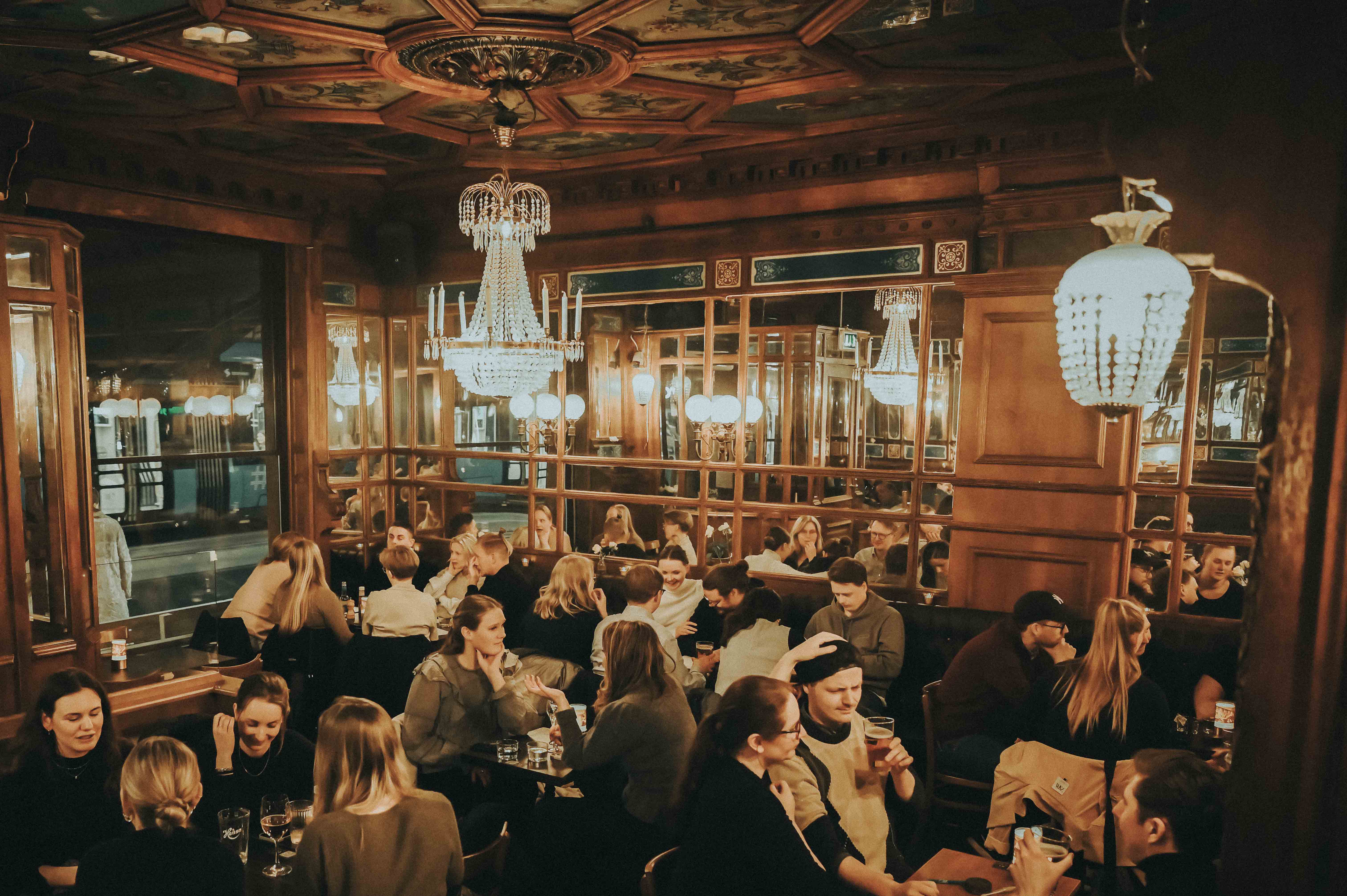 Mange mennesker sitter rundt bord p&aring; en m&oslash;rk bar med utsmykninger og lysekroner i taket